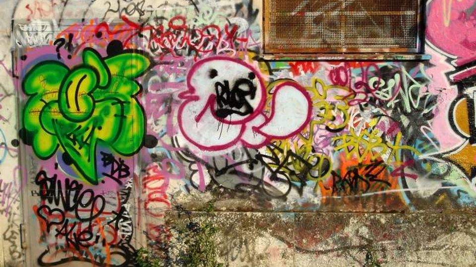 Parete esterna in cemento ricoperta da numerosi graffiti e scritte multicolori, con un disegno verde e una figura stilizzata rosa al centro. In alto a destra è presente una grata rettangolare su una finestra o presa d’aria, con vegetazione bassa alla base del muro.