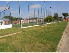 Campo sportivo recintato con alte reti metalliche e pali per l’illuminazione, affiancato da un prato con piccoli alberi. Sullo sfondo si vedono edifici bassi e una strada pedonale lungo il bordo destro.