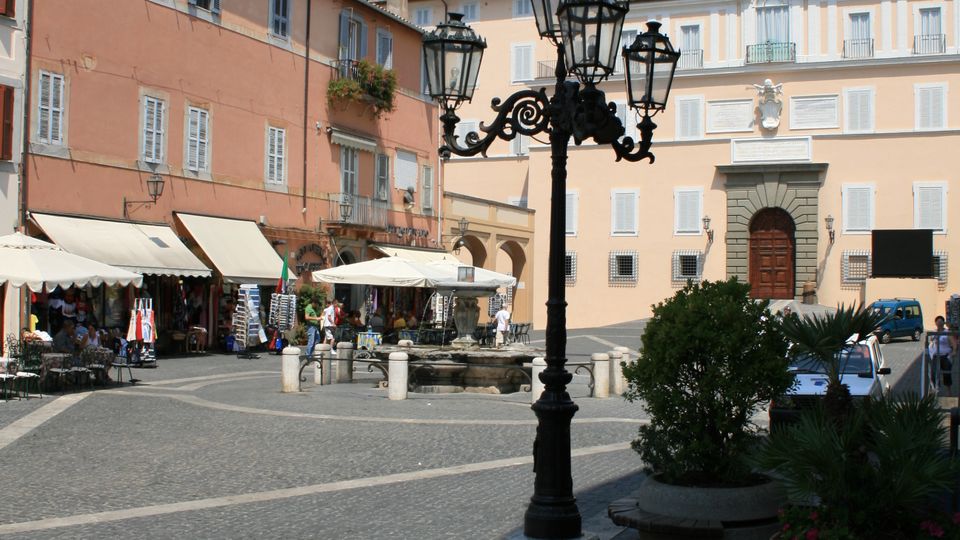 Piazza lastricata in un centro storico, con palazzi color pastello a più piani e un edificio istituzionale sullo sfondo con portale in pietra e piccolo frontone con orologio. In primo piano un lampione in ferro battuto, ai lati tende e bancarelle, ombrelloni e fioriere lungo il bordo della piazza.