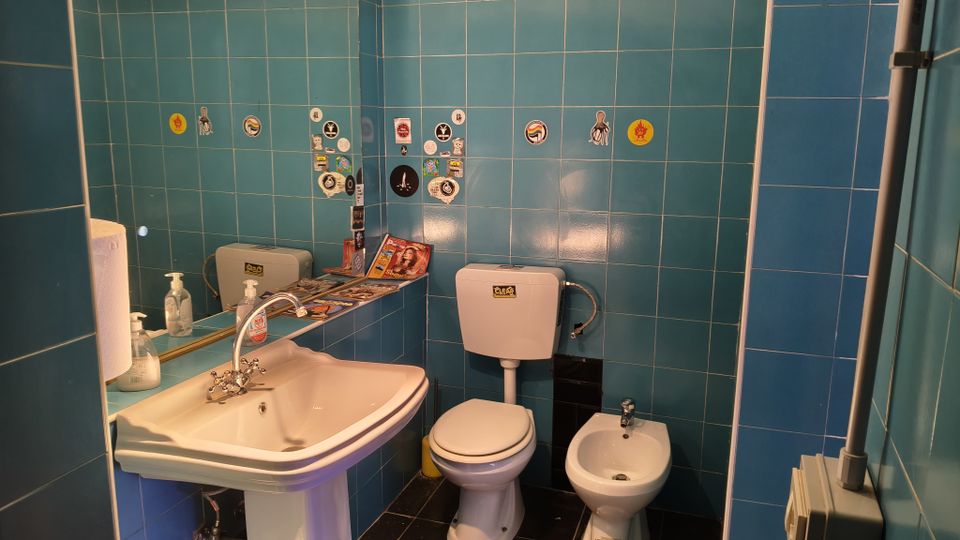 Piccolo bagno con pareti rivestite di piastrelle blu, lavabo bianco con specchio e dispenser di sapone. Sono presenti WC con cassetta esterna e bidet, con adesivi decorativi e una mensola con riviste sullo sfondo.