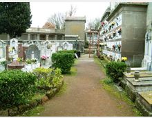 Viale di un cimitero con sentiero in ghiaia, bordato da siepi basse e tombe in pietra con lapidi e fiori. A destra si vede un muro di loculi sovrapposti con decorazioni floreali, con edifici funerari sullo sfondo.