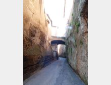 Vicolo stretto e in pendenza tra alte pareti di roccia e muratura, con strada asfaltata che curva in fondo. Un piccolo arco in pietra collega gli edifici sopra la via e sulle pareti si notano segni di erosione e vegetazione.