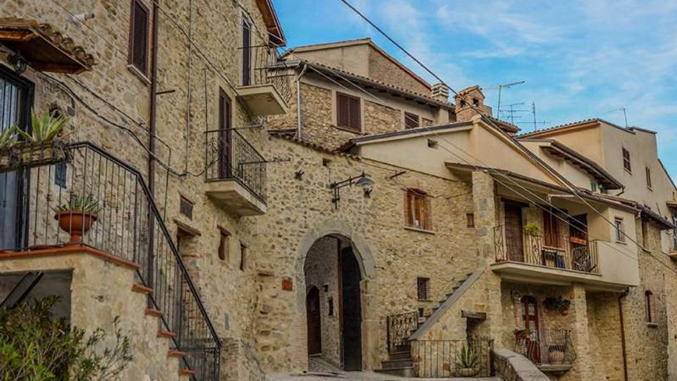 Vicolo acciottolato in un borgo storico con edifici in pietra a più piani, balconi e scale esterne con ringhiere in ferro battuto. Al centro si vede un arco d’ingresso e, sullo sfondo, tetti in coppi e cielo azzurro.