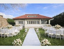 Giardino allestito per un ricevimento con tavoli rotondi coperti da tovaglie bianche e sedie chiare, disposti ai lati di una passerella centrale decorata con petali e lanterne. Sullo sfondo si vede una villa a un piano con tetto in tegole rosse e grandi finestre, circondata da mura in pietra e vegetazione.