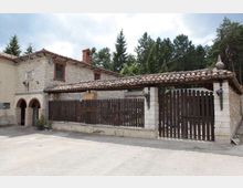 Edificio rurale in pietra a due piani con tetto in tegole e piccola loggia ad archi, affacciato su un cortile pavimentato. Davanti si vede una lunga recinzione e un grande cancello in legno con copertura in tegole, con alberi alti sullo sfondo.