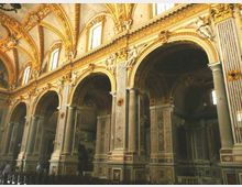 Interno di una chiesa con grandi arcate a tutto sesto sostenute da colonne e pilastri decorati, con stucchi dorati e statue lungo la cornice superiore. Pareti e soffitto presentano ricche decorazioni pittoriche e motivi ornamentali, illuminati da finestre alte.