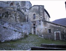 Antico complesso in pietra con edifici addossati a un pendio, mura in laterizio e una scalinata che conduce a un portale centrale. In primo piano si vede una piccola piazza erbosa con muretti e, sullo sfondo, il profilo delle colline.