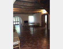 Ampio soggiorno rustico con pavimento in cotto, soffitto con travi in legno a vista e pareti bianche. Al centro c’è un tavolo, con divanetti in rattan, una finestra con tenda e una grande porta ad arco vetrata che dà sull’esterno.