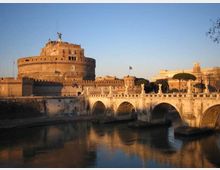 Veduta di Castel Sant’Angelo, fortezza cilindrica in muratura, con il Ponte Sant’Angelo in primo piano caratterizzato da archi in pietra e statue lungo la balaustra. Il ponte attraversa il fiume Tevere, con riflessi sull’acqua e cielo sereno sullo sfondo.