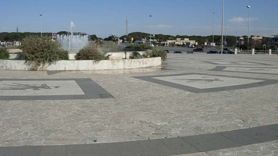 Ampia piazza pavimentata in pietra con grandi riquadri decorativi sul suolo e una fontana circolare al centro, circondata da aiuole. Sullo sfondo si vedono una strada con auto, lampioni alti e una linea di alberi sotto un cielo sereno.