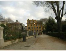 Viale pedonale in un parco urbano con sentiero in ghiaia e pavé, fiancheggiato da siepi, alberi e una recinzione in ferro battuto su muretto. Sullo sfondo si vede un edificio a più piani color ocra e lampioni lungo il percorso.