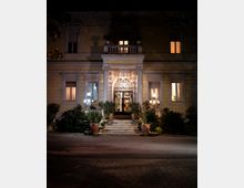 Facciata notturna di una villa storica con ingresso centrale ad arco, portone in ferro battuto e scalinate in pietra. Colonne, balcone sopra l’entrata, finestre illuminate e giardino con piante in vaso e lampioni ai lati incorniciano l’accesso.