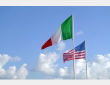 Due pennoni con la bandiera italiana e la bandiera degli Stati Uniti che sventolano all’aperto contro un cielo azzurro con nuvole bianche.
