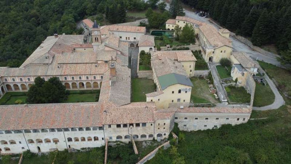 Veduta aerea di un grande complesso monastico o abbaziale con edifici in pietra e intonaco, tetti in coppi e cortili interni, con una chiesa e un campanile al centro. Il complesso è circondato da boschi e prati, con una strada di accesso sul lato destro.