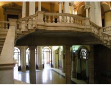 Interno di un edificio storico con grande scala monumentale e ballatoio in pietra con balaustra, sostenuti da colonne. Al livello inferiore si vedono archi e alte finestre ad arco che illuminano un ampio atrio.