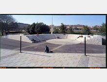 Anfiteatro all’aperto in pietra chiara con gradoni semicircolari e area centrale pavimentata, inserito in una piazza con lampioni e vialetti. Sullo sfondo si vedono alberi e edifici urbani sotto un cielo sereno.