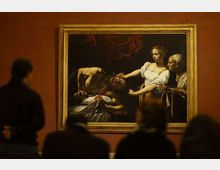 Sala museale con pareti color ocra e un grande dipinto incorniciato in oro appeso al centro. Davanti al quadro si intravede il profilo sfocato di visitatori, mentre l’opera raffigura una scena drammatica con figure illuminate su sfondo scuro.