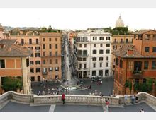 Veduta dall’alto di una piazza urbana con una fontana centrale e una lunga strada rettilinea che si estende tra palazzi storici dai toni ocra e bianco. In primo piano si vede una balaustra in pietra e, sullo sfondo, i tetti della città con una grande cupola visibile in lontananza.