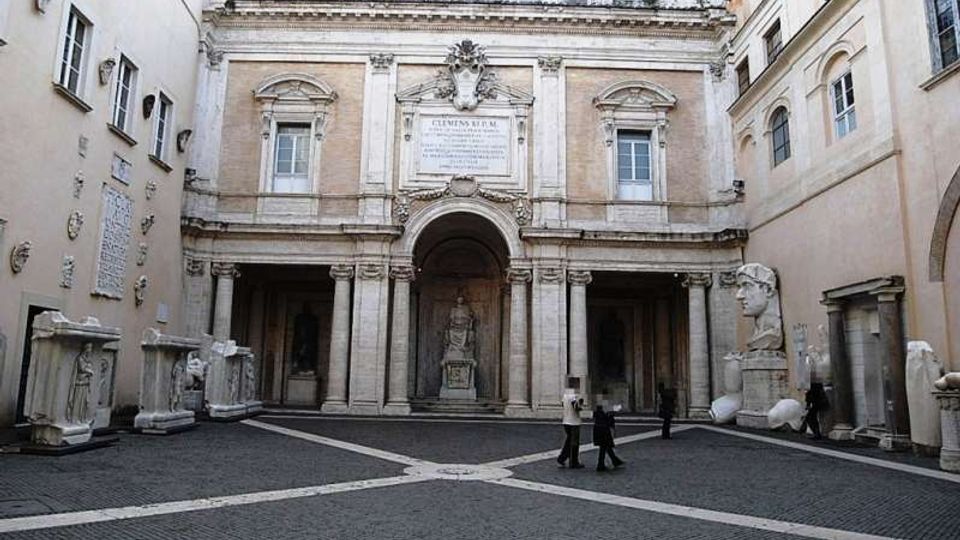 Cortile interno di un complesso storico con pavimentazione in selciato e facciata monumentale in pietra con grande arco centrale e porticato. Ai lati sono disposte statue e frammenti scultorei, tra cui un grande busto, con edifici chiari che chiudono lo spazio su tre lati.