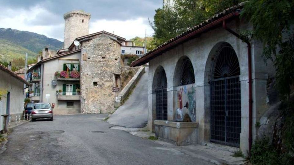 Strada di un borgo montano con edifici in pietra e una torre cilindrica sullo sfondo. A destra si vede un lungo edificio con portico ad archi e grate metalliche, mentre a sinistra ci sono case con balconi e la strada in lieve curva.
