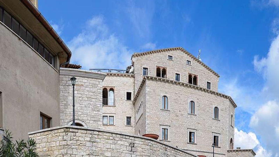 Edificio storico in pietra chiara, simile a un castello o monastero, con più livelli e finestre ad arco, visto dal basso dietro un alto muro in pietra. Sullo sfondo si vede il cielo azzurro con nuvole e un sentiero erboso lungo il perimetro.