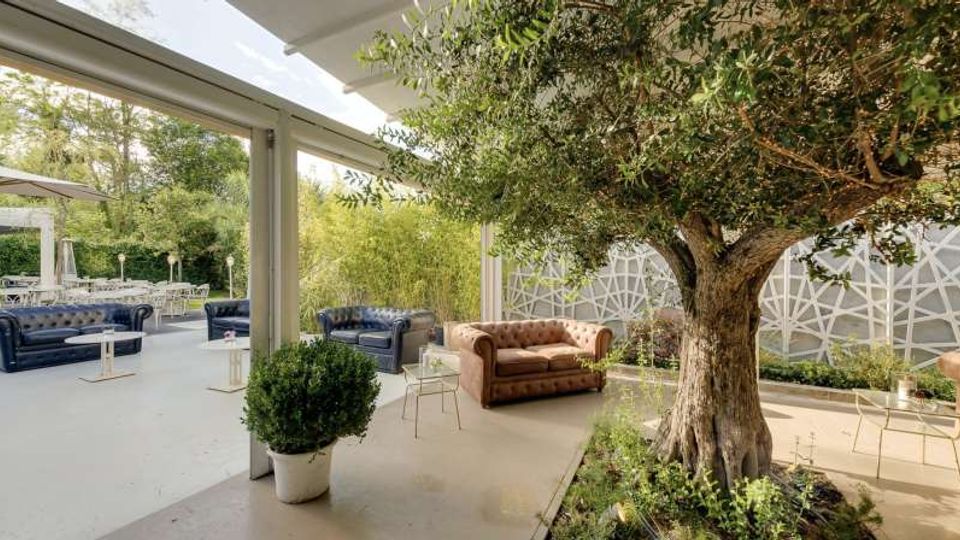 Terrazza coperta con pavimento chiaro, arredata con divani in pelle stile Chesterfield e tavolini bassi, affacciata su un’area verde. In primo piano un grande albero in una aiuola, con parete di fondo decorata da pannelli geometrici e vegetazione lungo il perimetro.