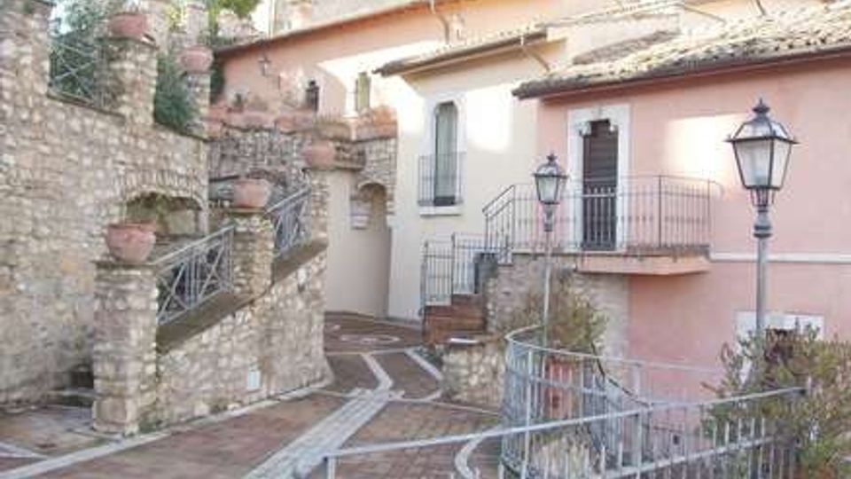 Cortile in pietra di un edificio storico con facciata rosa, scale e terrazze con ringhiere in ferro battuto e vasi in terracotta lungo i muri. Pavimentazione a mosaico e lampioni in stile classico, con strutture in pietra e tetti in tegole sullo sfondo.