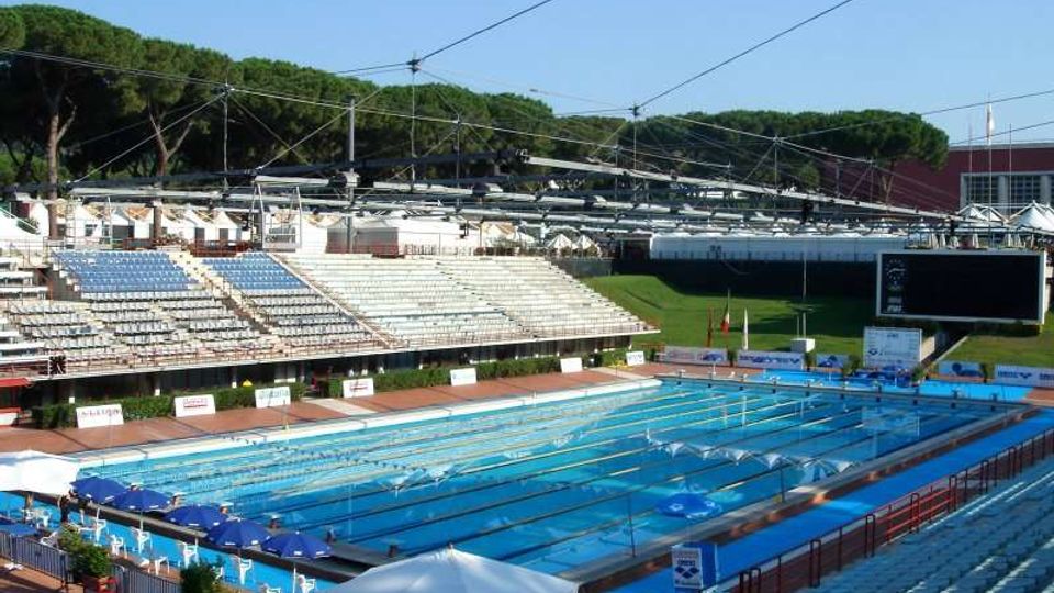 Piscina olimpionica all’aperto con corsie delimitate e trampolini, circondata da tribune con gradinate e sedute. Sullo sfondo si vedono una grande schermata elettronica, strutture metalliche con cavi e un’area verde con alberi.