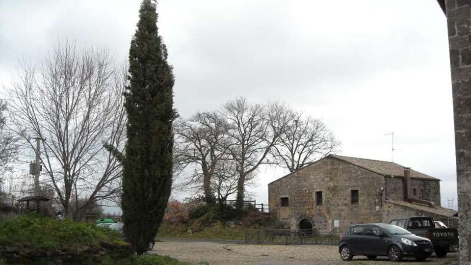 Piazzale lastricato in pietra con un alto cipresso e alberi spogli, affacciato su un edificio rurale in pietra con tetto a falde. Sullo sfondo cielo nuvoloso e alcune auto parcheggiate lungo il bordo della piazza.