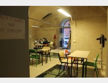 Interno di una sala studio in un edificio con pareti in intonaco grezzo e soffitto a volta, illuminata da lampade lineari. Sono presenti tavoli con sedie di colori diversi, un pavimento in piastrelle con inserti in vetro e un grande portale ad arco che si apre verso l’esterno.