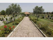 Viale pavimentato in un cimitero, fiancheggiato da aiuole fiorite e alberi, con file di lapidi e monumenti funerari su entrambi i lati. In fondo al percorso si vede una piccola struttura con tetto in tegole.