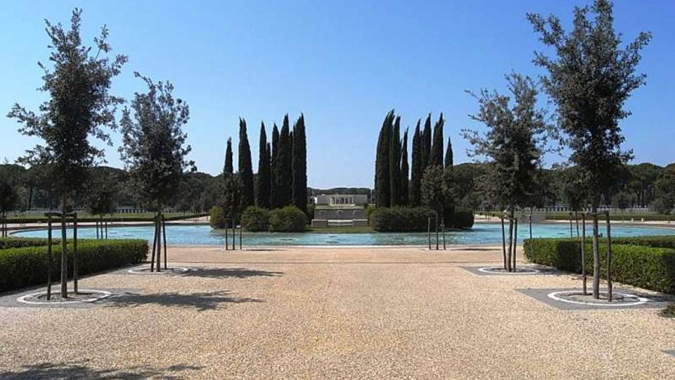 Viale pedonale in ghiaia in un parco formale, fiancheggiato da giovani alberi e siepi basse, che conduce a una grande vasca d’acqua con un’isola di cipressi. Sullo sfondo si vede un edificio basso e simmetrico oltre la fontana, con alberi e bosco tutt’intorno sotto un cielo sereno.