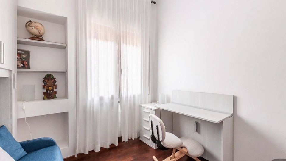 Piccolo studio domestico con pareti bianche, pavimento in legno scuro e finestra con tende leggere. A destra c’è una scrivania bianca con cassettiera e sedia ergonomica, mentre a sinistra si vedono mensole a incasso e un divano blu.