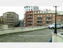 Terrazza urbana in cemento con pavimentazione a grandi lastre e parapetto basso, affacciata su una strada tra alti edifici residenziali in mattoni con balconi. In primo piano si vedono un lampione e un’auto parcheggiata vicino al bordo della terrazza.