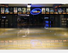 Ingresso di un cinema con pavimento lucido in piastrelle chiare e riflessi, corridoio centrale illuminato e insegna al neon “UCI CINECITTÀ” sopra l’entrata. Lungo la parete superiore sono visibili vetrine illuminate con locandine di film e, ai lati, chioschi automatici e bacheche informative.