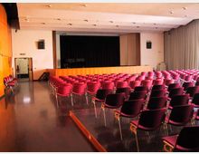 Sala auditorium con file di sedie rosse e nere rivolte verso un palco rialzato con sipario scuro. Pareti chiare con pannelli in legno, illuminazione a soffitto e uscita laterale sulla sinistra.