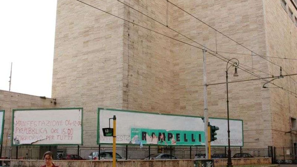 Grande edificio urbano in pietra chiara con volumi squadrati e poche finestre, sormontato da antenne, visto dall’angolo di una strada con binari del tram e cavi aerei. In primo piano ci sono un incrocio con semafori, una recinzione e pannelli pubblicitari lungo il perimetro dell’edificio.
