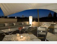Terrazza panoramica su un tetto con tavoli apparecchiati e divani, illuminata da lampade e candele sotto una grande copertura bianca. Sullo sfondo si vede lo skyline urbano notturno con cupole e edifici storici.