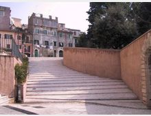 Ampia scalinata in pietra che sale tra muri intonacati color ocra, con lampione e ringhiera sul lato sinistro. In cima si affacciano edifici residenziali alti e vissuti con persiane e archi al piano terra, accanto a grandi alberi.
