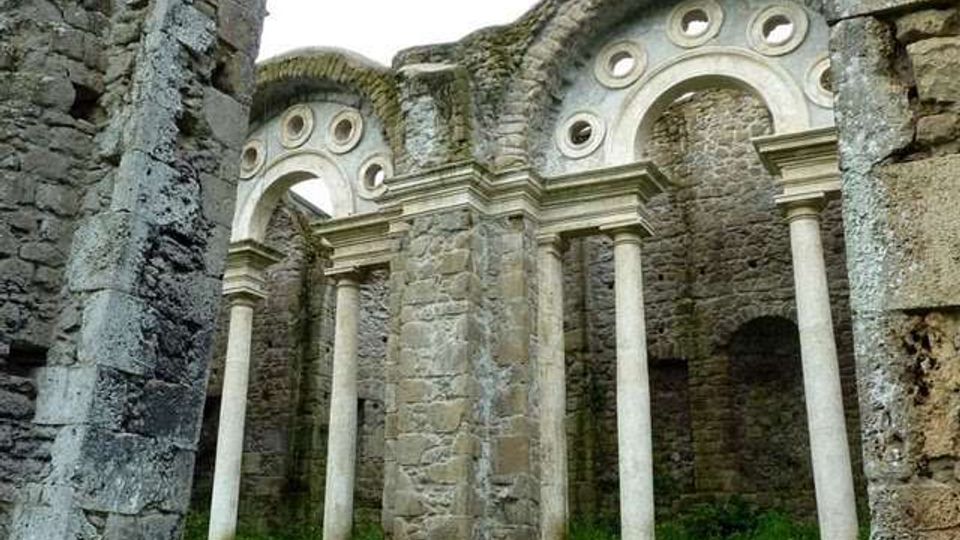 Rovine di una chiesa in pietra con abside semicircolare, archi decorati da oculi circolari e una serie di colonne slanciate lungo le pareti. In primo piano si vedono murature in pietra grezza e un prato che cresce all’interno della struttura priva di tetto.
