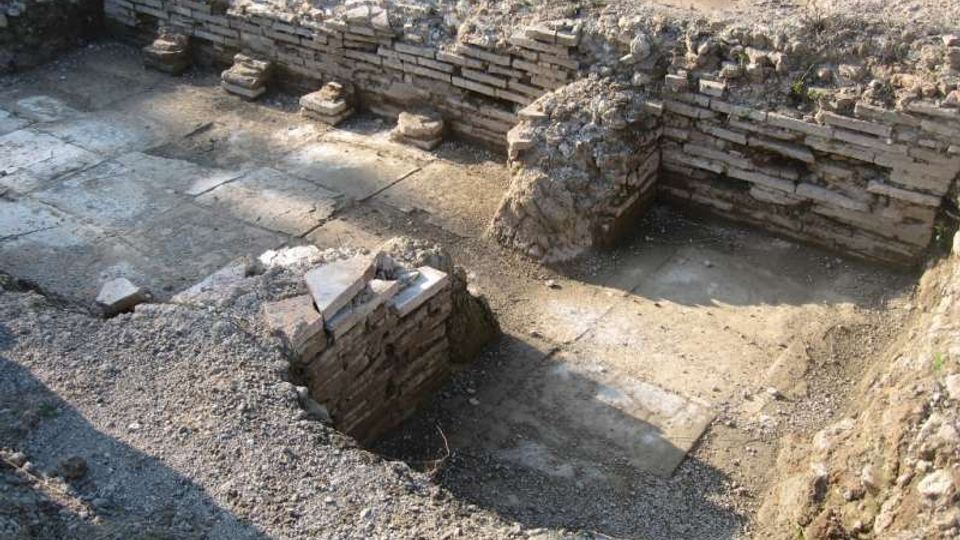 Scavo archeologico con una struttura rettangolare interrata, pavimentata con grandi lastre di pietra e delimitata da muretti in mattoni e pietra. Si vedono piccoli basamenti o gradini lungo il lato e terreno smosso tutt’intorno.