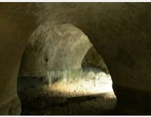 Interno di una piccola galleria sotterranea in pietra con volta ad arco, pareti grezze e pavimento in terra e ghiaia. Una zona illuminata in fondo mette in evidenza depositi e macchie di umidità lungo le pareti.