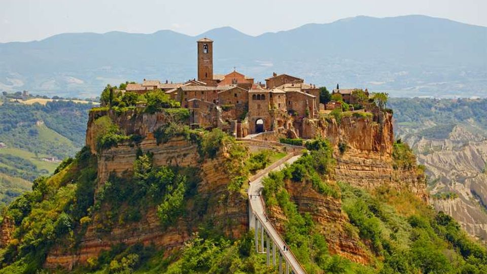 Borgo storico in pietra arroccato su una rupe di tufo, con campanile e case compatte attorno a un ingresso ad arco. Un lungo ponte pedonale sopraelevato collega il paese al crinale, con colline verdi e montagne sullo sfondo.