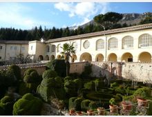 Cortile di un complesso storico con porticato ad archi e grandi finestre ad arco, affacciato su un giardino formale con siepi potate e vasi in terracotta. Sullo sfondo si vedono alberi, montagne rocciose e cielo parzialmente nuvoloso.