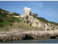 Castello in pietra con torre cilindrica e merli, arroccato su una scogliera rocciosa sopra il mare. Il pendio è coperto di vegetazione mediterranea e muri di contenimento, con un edificio adiacente e cielo sereno sullo sfondo.