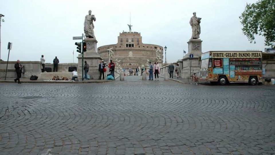 Piazza lastricata in sampietrini con accesso a un ponte, fiancheggiato da due statue su alti piedistalli. Sullo sfondo si vede Castel Sant’Angelo, mentre a destra è parcheggiato un chiosco ambulante per cibo con insegna.