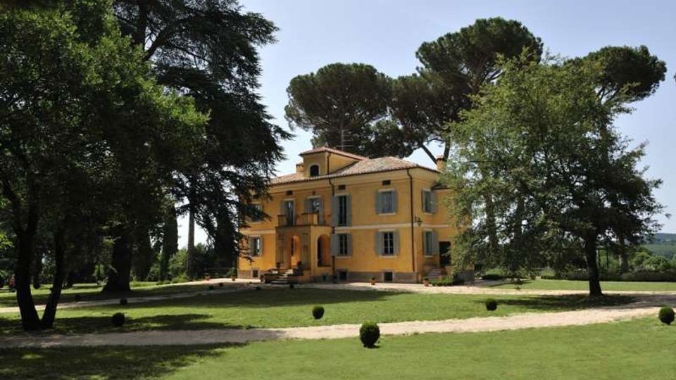 Villa storica color ocra a due piani con tetto in coppi e persiane, al centro di un ampio parco. Davanti all’edificio si vedono prati curati, vialetti in ghiaia e grandi alberi ad alto fusto che incorniciano la facciata.