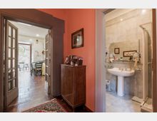 Corridoio interno con pareti rosa e pavimento in legno, con mobile in legno e porta a vetri che conduce a una cucina-sala da pranzo con accesso a un balcone. Sulla destra si apre un bagno piastrellato in beige con lavabo su colonna, specchio e box doccia.