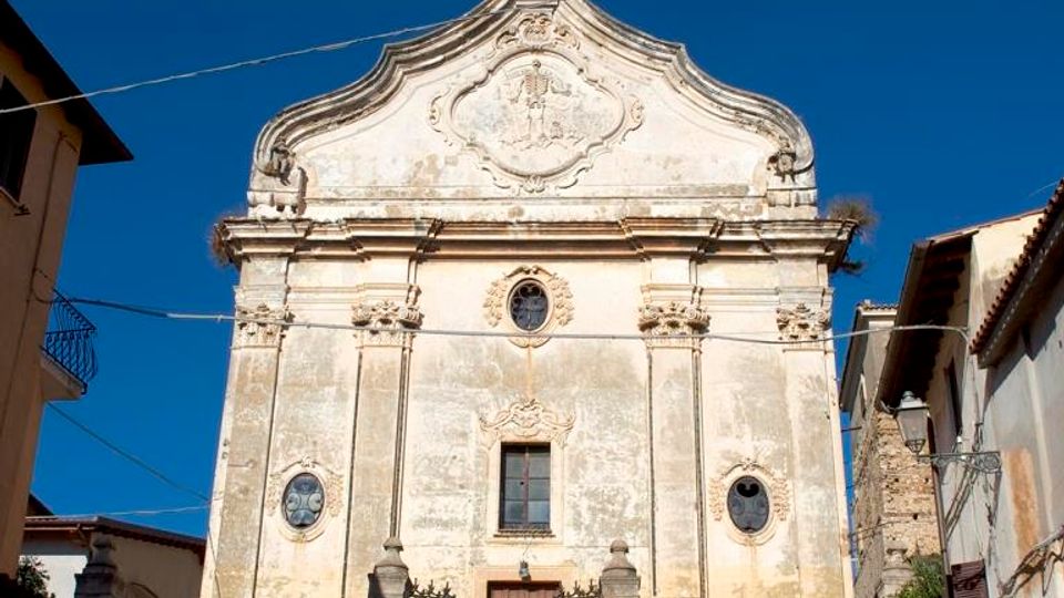 Facciata di una chiesa storica in stile barocco, con frontone curvilineo decorato, finestre ovali laterali e ingresso centrale sopra una scalinata in pietra con cancellata in ferro. La chiesa è affiancata da edifici residenziali e attraversata da cavi sospesi nel cielo azzurro.