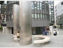 Piazza urbana moderna con due edifici cilindrici in vetro e metallo e una grande colonna centrale rivestita in acciaio. Nell’area pedonale ci sono panchine curve in cemento e facciate vetrate che circondano lo spazio.